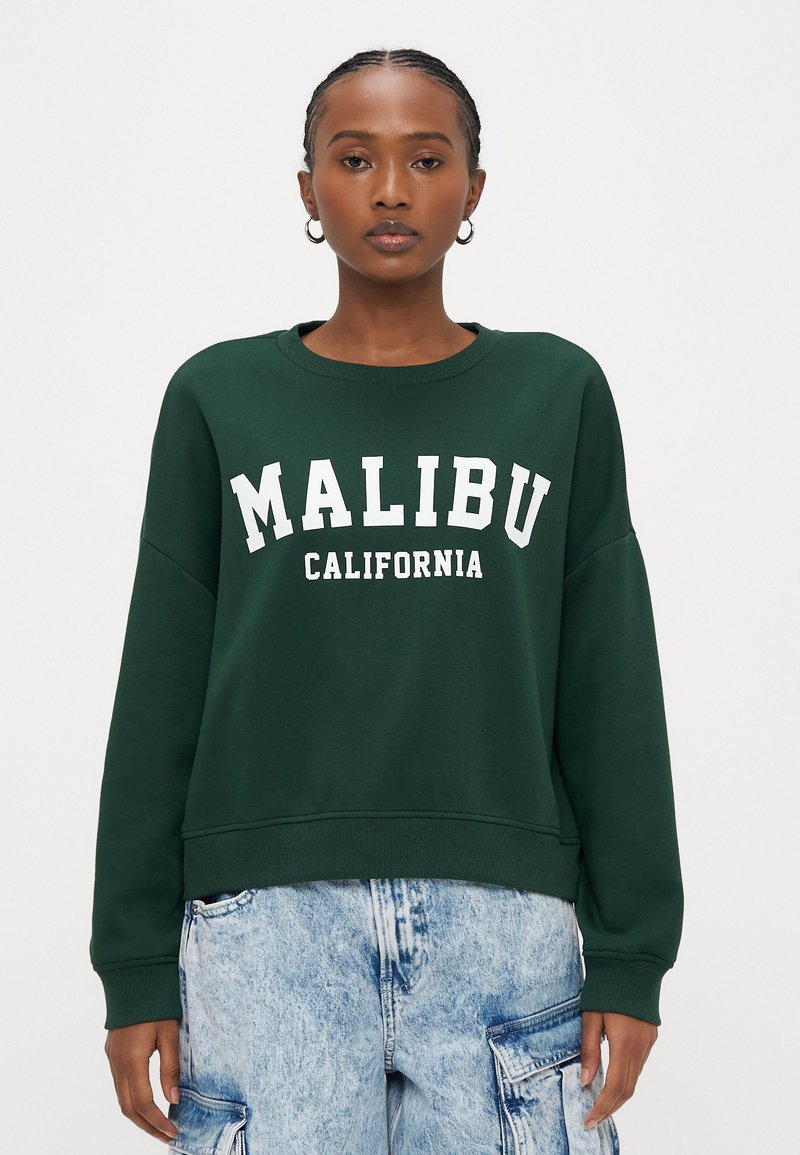 Grüner Pullover mit dem weißen Schriftzug "MALIBU CALIFORNIA". Verfügt über einen Rundhalsausschnitt und eine lockere Passform, kombiniert mit hellblauen Denim-Cargohosen.