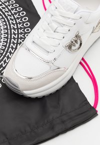 Fehér sneaker texturált hálós anyaggal és velúr kiegészítőkkel, ezüst hardware részletekkel és rózsaszín fűzőkkel, elhelyezve egy fekete húzós zacskón, amelyen logó található.
