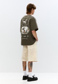 T-shirt oversized verde oliva con stampe grafiche sulla schiena, abbinata a pantaloni corti beige chiaro tie-dye e scarpe nere con lacci.