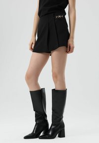 Zwarte geplooide shorts met een decoratieve gouden kettingaccent bij de tailleband, gecombineerd met kniehoge zwarte leren laarzen met vierkante hakken.