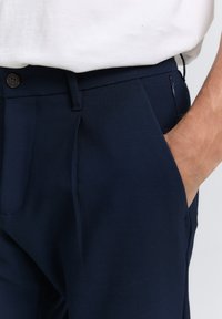 Pantalones azul marino de tela suave con un diseño de frente plano, que cuentan con un cierre de botón y detalles en los bolsillos laterales.