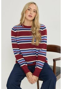 Maroon gestreepte trui met blauwe en crèmekleurige banden, ronde hals, geribbelde manchetten en zoom. Gede gedragen met losse donkere spijkerbroek, zittend op een houten stoel.