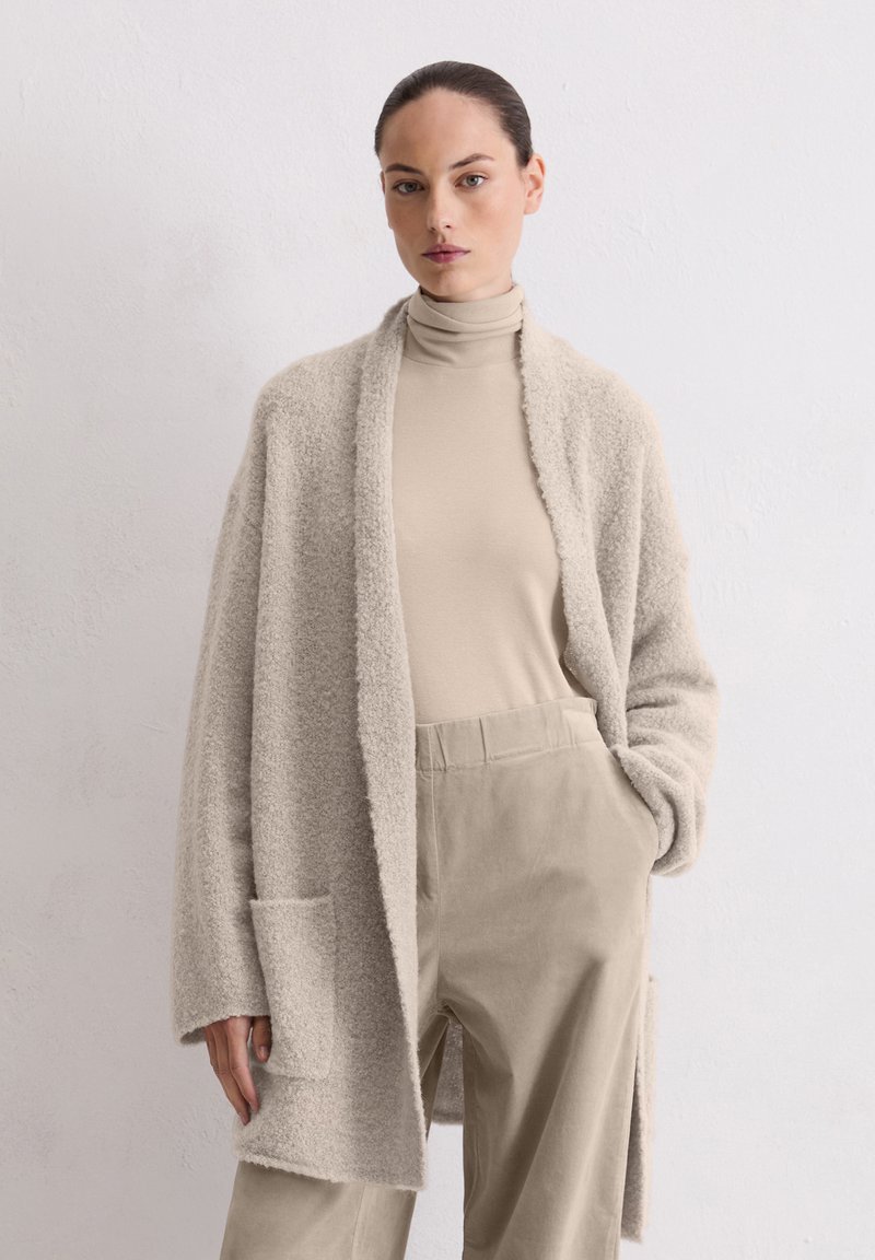 Licht beige fuzzy cardigan met een open voorkant, voorzien van een grote zak. Draag het over een bijpassende turtleneck en zachte beige broek.