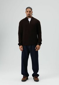 Bruine gebreide trui met ritssluiting en hoge kraag, gecombineerd met losse blauwe spijkerbroeken en bruine wandelschoenen. Eenvoudig en functioneel ontwerp.