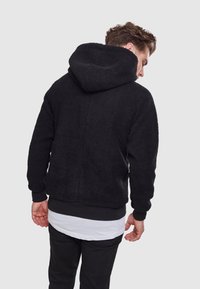 Giacca in pile nera con cappuccio, caratterizzata da un esterno testurizzato, polsini elasticizzati e una silhouette arrotondata. Strati visibili sotto la giacca.