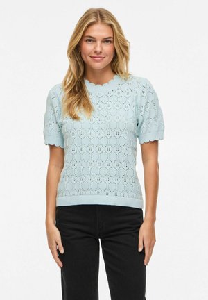 Femme blonde portant un haut en maille bleu clair à manches courtes avec un ourlet festonné et un pantalon noir, debout devant un fond blanc.