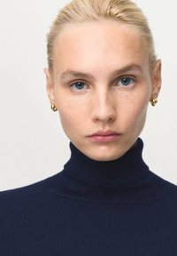Blonde vrouw met blauwe ogen en lichte sproeten, die gouden oorringen en een marineblauwe coltrui draagt, recht vooruit kijkend tegen een neutrale achtergrond.
