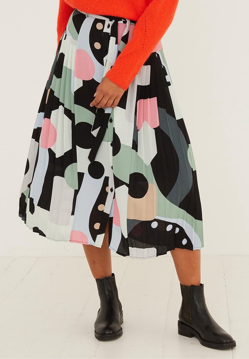 Persoon draagt een geplooide midi-rok met abstracte print in pasteltinten, zwarte enkellaarzen en een oranje trui met lange mouwen, staand binnen.