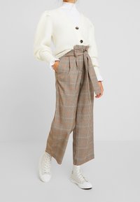 Cardigan en tricot crème avec col en V et deux boutons, associé à un pantalon à carreaux marron taille haute avec ceinture à nouer et baskets blanches montantes à la cheville.