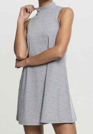 Mouwloze grijze A-lijn jurk met een opstaande kraag, gedragen door een persoon die de kraag met één hand vasthoudt en de andere arm gekruist heeft.