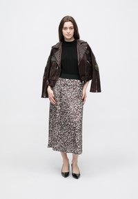 Kobieta ubrana w ciemną, ćwiekowaną skórzaną kurtkę na czarnym topie i długą spódnicę w panterkę, na czarnych, szpiczastych obcasach, stojąca na białym tle.