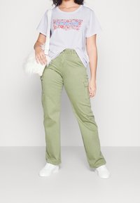 T-shirt gris clair avec un motif floral, associée à un pantalon cargo vert olive avec poches latérales. Le modèle tient un sac en fourrure blanche et porte des baskets blanches.