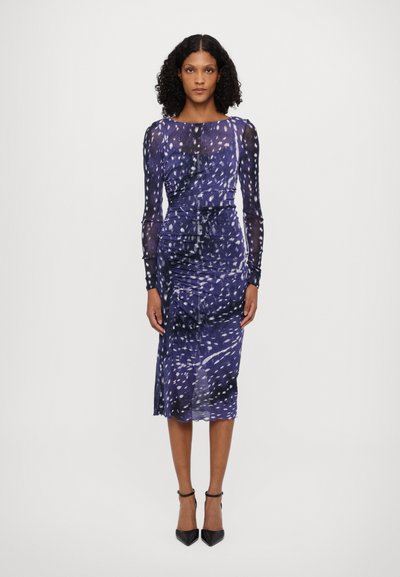 Langes, lila Kleid mit weißen abstrakten Mustern; geraffter Stoff an der Taille, knielanger Saum, kombiniert mit schwarzen Ankle-Strap-Absätzen.