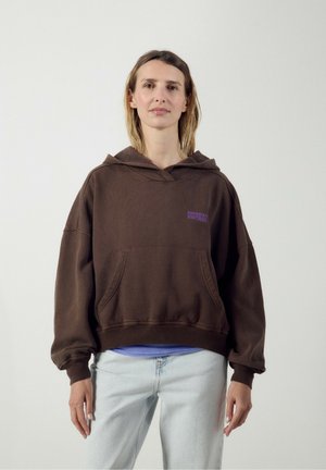 Femme aux cheveux mi-longs portant un sweat à capuche marron oversize avec le texte "American Vintage", jean bleu clair, debout devant un fond uni.