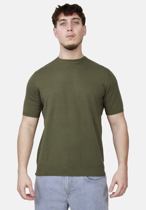 Giovane uomo con capelli corti e ricci, che indossa una camicia verde oliva a maniche corte e jeans azzurri, in piedi davanti a uno sfondo semplice.