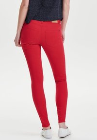 Jean skinny rouge avec une texture lisse, deux poches arrière et une coupe ajustée. Des chaussures blanches avec un détail texturé complètent le look.
