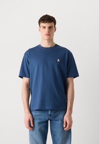 Blå kortærmet T-shirt lavet af bomuld med en rund halsudskæring og et lille trekantet logo på venstre bryst. Denimjeans bæres nedenunder.