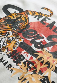 White fabric featuring a graphic design of a tiger in orange and black, surrounded by red accents and bold black text, with a textured appearance.