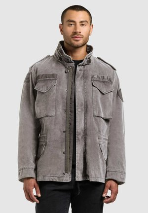 Mand iført en grå militærinspireret jakke med lynlås og store klaplommer samt sorte bukser, stående mod en ensfarvet baggrund.