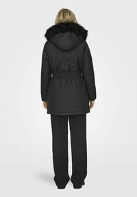 Svart parkas med hette av faux pels, innsvingt midje, lange ermer og rett snitt. Jevn tekstur og midtlengde design.