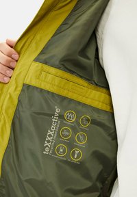 Grøn og gul vandtæt jakke med genanvendt membran, der har tapeforseglede sømme, åndbart materiale og ikoner, der detaljerer vandtætheden.