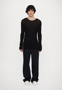 Chemise noire transparente à manches longues avec des bras ajustés et un col rond, associée à un jean bleu foncé ample et des baskets blanches.