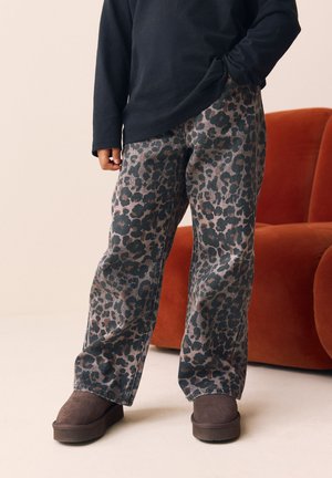 Niño con pantalones de estampado de leopardo, botas marrón oscuro y una camiseta negra de manga larga, de pie junto a un sillón naranja.