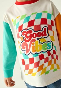 Camicia bianca a maniche lunghe con maniche turchesi e arancioni. Presenta un motivo a quadri colorato e la scritta "Good Vibes" in un font retro.
