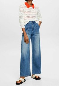 Weißer Strickpullover mit rotem Kragen, weite blaue Jeans und beigefarbene Sandalen mit schwarzen Sohlen. Erkennbar sind strukturierte Muster und Details mit lockerer Passform.