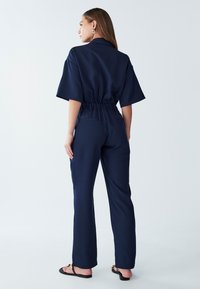 Mornaric jumpsuit s kratkimi rokavi, elastičnim pasom in ravnimi hlačami. Ima minimalisticen dizajn in gladko teksturo. Ni vidnih vzorcev ali dodatkov.