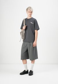 Graues T-Shirt mit weißem Text, lockere graue Shorts, weiße Socken und schwarze Slipper, kombiniert mit einer beigen Umhängetasche.