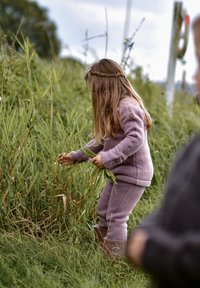 Barn i en lyse lilla fleecetrøje, der krøller sig sammen for at samle lang, grøn græs, med tekstureret stof og et flettet pandebånd synligt.