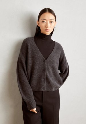 Mörkgrå stickad cardigan med V-ringning, åtta knappar framtill och vida ärmar, lagd över en svart polotröja.