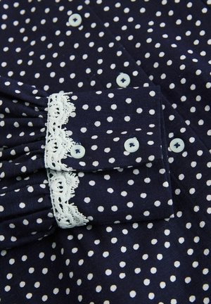 Navy blue blouse with white polka dots. Features a ruffled cuff with white lace trim and light blue buttons. Textured fabric visible.
