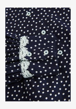 Marineblaue Bluse mit weißen Polka-Dots. Verfügt über eine geraffte Manschette mit weißer Spitzenverzierung und hellblauen Knöpfen. Strukturierter Stoff sichtbar.
