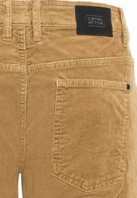 Pantalon en velours côtelé beige avec une poche arrière, des coutures apparentes et une étiquette noire avec le logo "camel active" sur la ceinture.