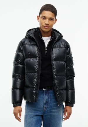 Mann trägt schwarze glänzende Steppjacke mit Kapuze, schwarzen Reißverschluss-Pullover, weißes Hemd und blaue Jeans vor weißem Hintergrund.
