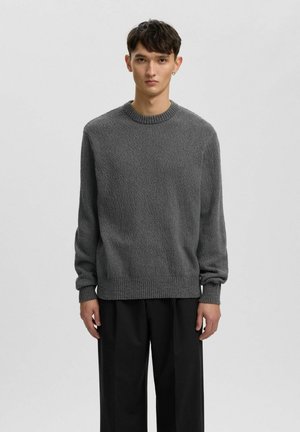 Ung mand står mod en ensfarvet baggrund iført en struktureret grå sweater og løse sorte bukser, vender fremad med et neutralt udtryk.