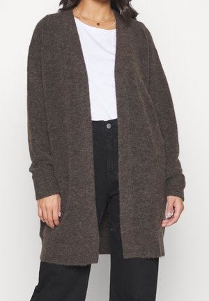 Femme portant un cardigan marron foncé ouvert sur le devant et en tissu pelucheux, par-dessus une chemise blanche et un pantalon noir taille haute, visible du cou jusqu’aux hanches.