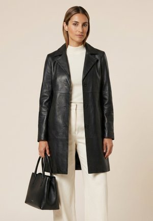 Vrouw met een zwarte leren jas over een wit topje en broek, houdt een zwarte leren handtas vast tegen een beige achtergrond.