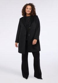 Černý vlněný kabát s klopami, dvěma předními knoflíky a bočními kapsami. Látka má hladkou texturu a je nošena přes černé kalhoty.