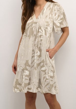 Vrouw draagt een losse beige en witte jurk met bloemenpatroon, korte mouwen en een V-hals, staand met één hand in de zak.