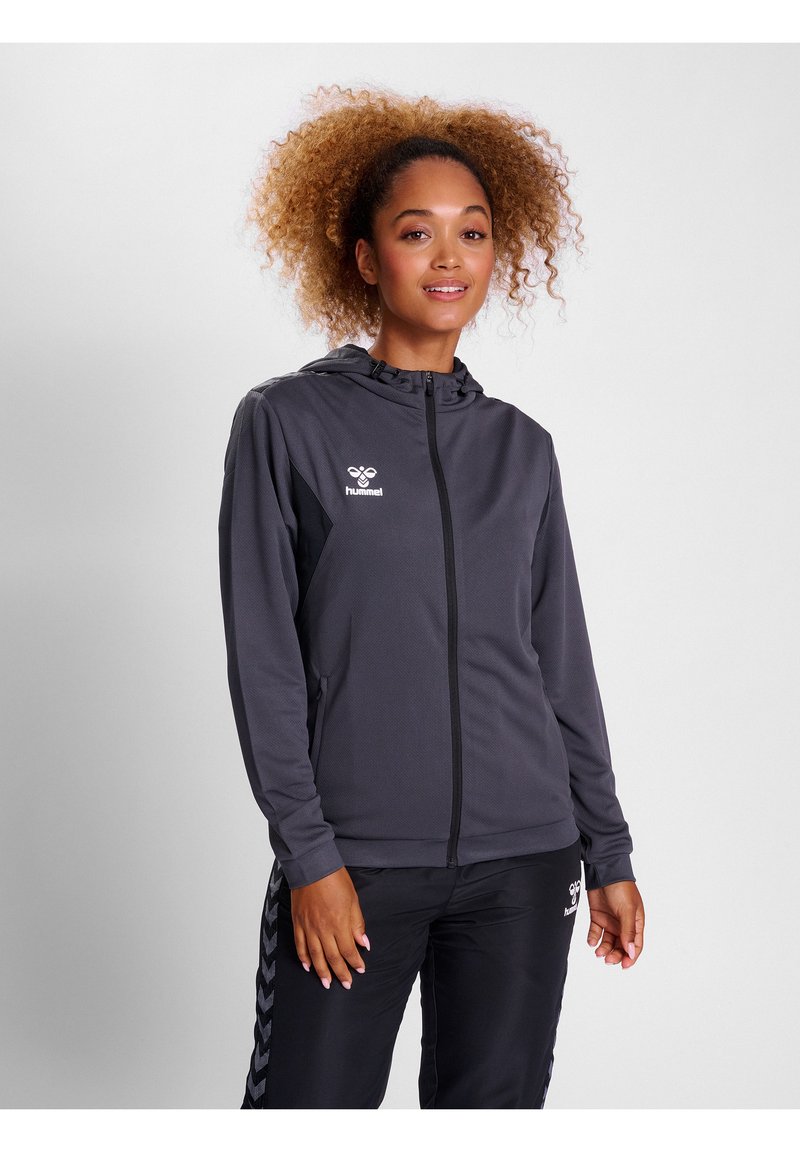 Grau Zip-up Kapuzenpullover mit Kapuze, mit einem Logo auf der Brust, langen Ärmeln und strukturiertem Stoff. Getragen mit schwarzen Hosen und einer lockeren Passform.
