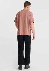 Man in een losse roze korte mouwen shirt, zwarte broek en zwarte schoenen, staand met de rug naar de camera tegen een egale lichte achtergrond.