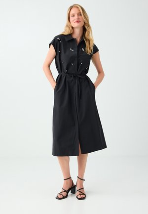 Femme portant une robe chemise midi noire à manches courtes, détails en sequins, taille nouée et sandales noires à talons blocs et brides.