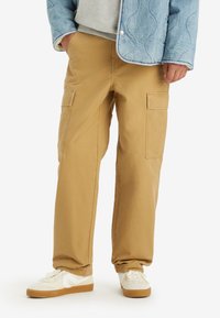Beige cargo pants with multiple side pockets, relaxed fit, and soft cotton fabric, paired with white sneakers and a light blue quilted jacket.