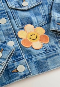Jeansjacke in blauer Waschung mit silbernen Druckknöpfen und einem bestickten Blumenpatch in Orange und Gelb mit einem lächelnden Gesicht.