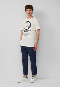 Witte graphic t-shirt met blauwe tekst "PACIFIC CALIFORNIA" en een cirkelontwerp, gecombineerd met marineblauwe tapered pants en witte sneakers.