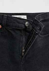 Gros plan sur un jean en denim noir avec une fermeture éclair partiellement ouverte et un bouton en métal à la taille montrant une étiquette blanche de taille à l'intérieur.