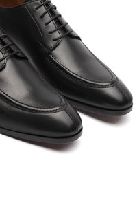 Scarpe in pelle nera con finitura liscia, punta affusolata e cuciture decorative. Il design include lacci e una suola in gomma minimalista.